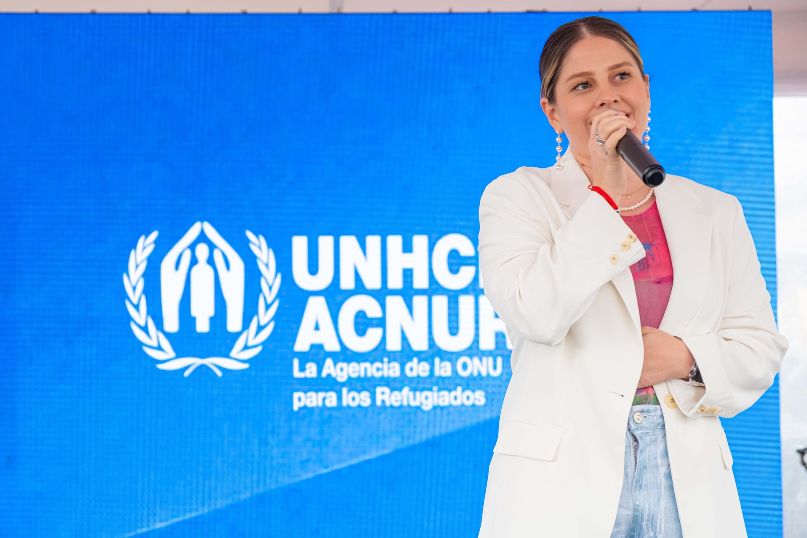 Una mujer que viste ropa blanca, sostiene un micrófono. Detrás de ella un fondo azul con el logo de ACNUR en blanco