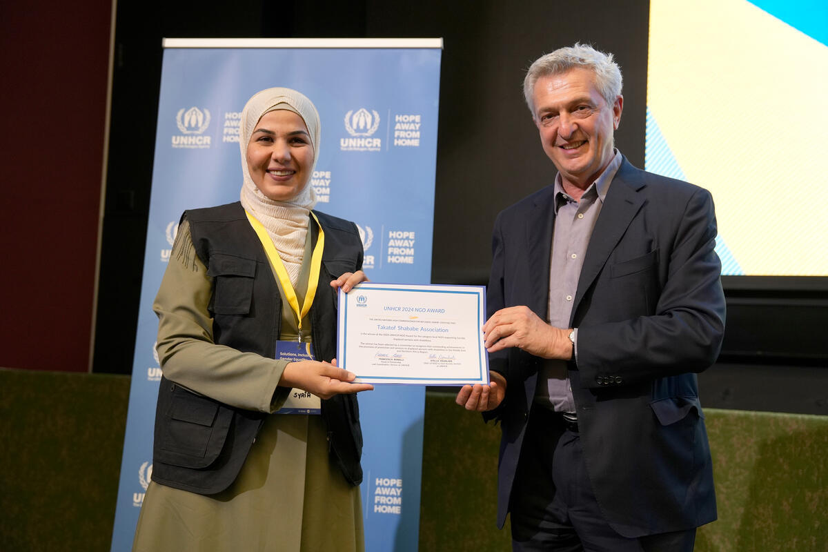 Una mujer, que lleva puesto un hijab color blanco, sonríe y muestra un certificado junto a un hombre cano