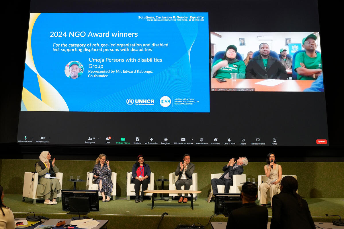  Un panel de seis personas aplaude y observa la pantalla detrás suyo, donde se proyecta la imagen de un líder refugiado que no pudo asistir presencialmente al evento