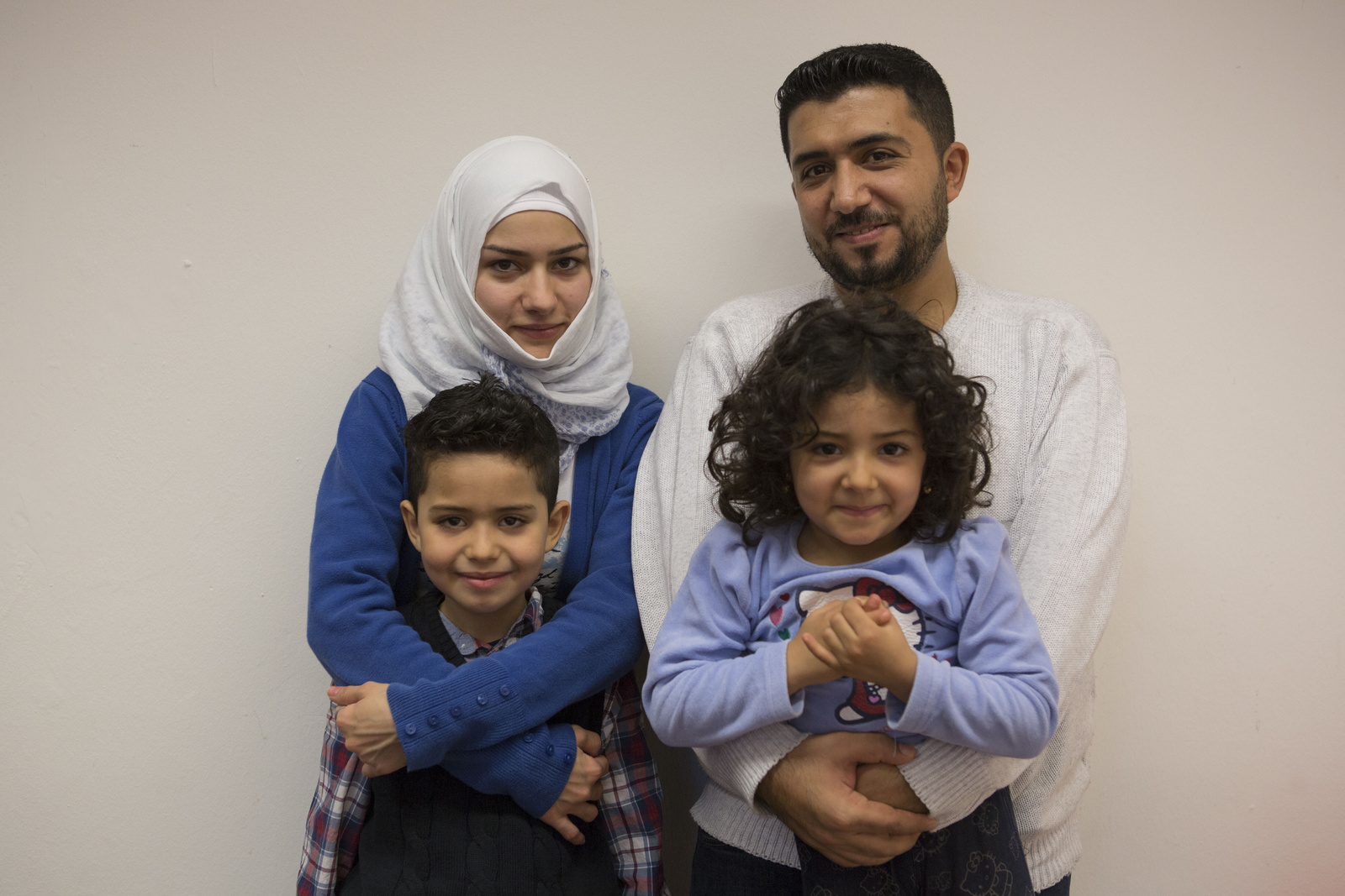 Una familia de cuatro personas (madre, padre, hija e hijo) sonría a la cámara. Ella, que lleva puesto un jiyba de color blanco, abraza a su hijo; él abraza a la niña, que tiene el cabello rizado.