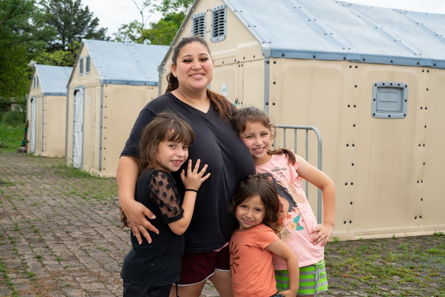 Una mujer con tres niñas posan para la foto delante de unas casitas prefabricadas.