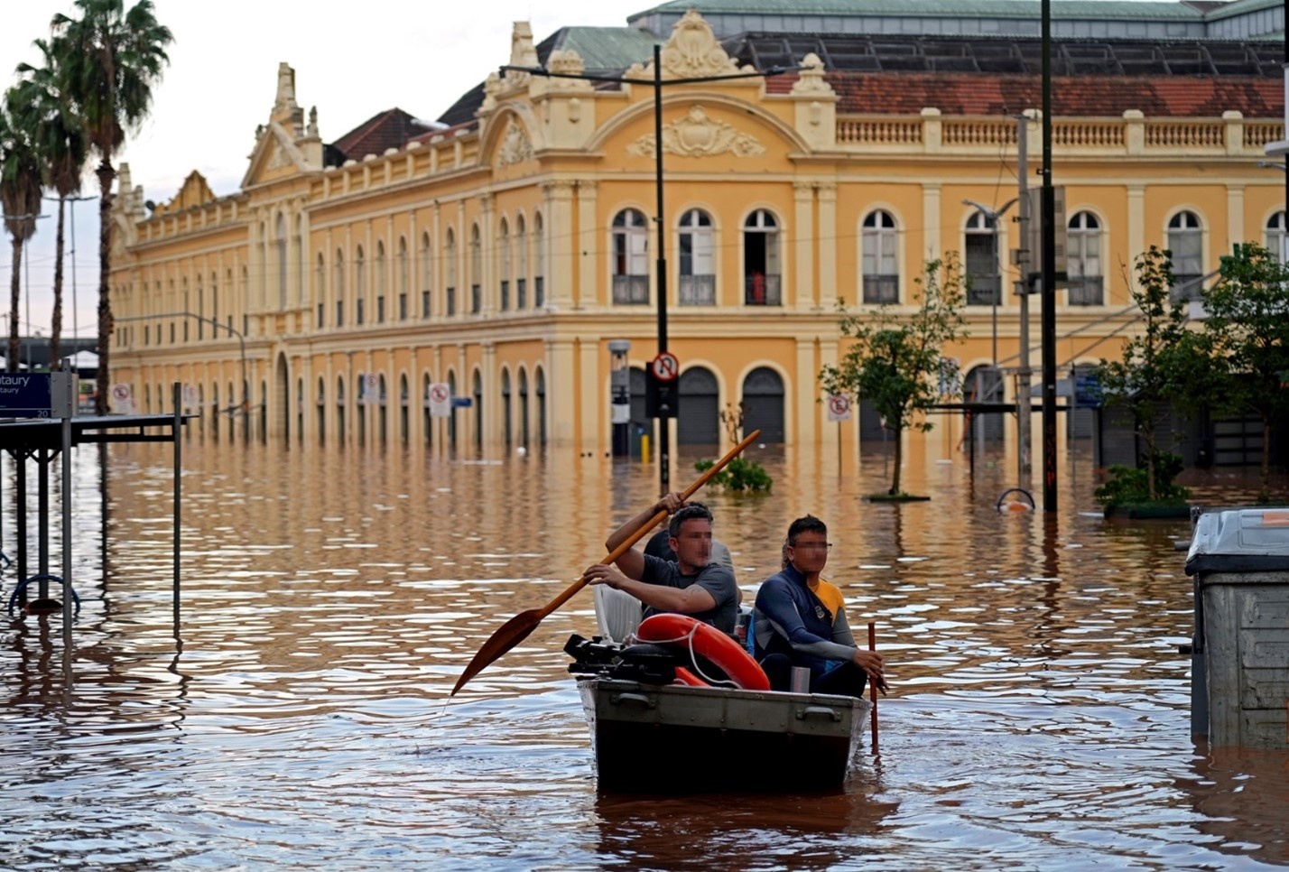 Dos personas se desplazan en un bote en una calle complemtamente inundada en un centro urbano. A sus espaldas un edificio en estilo clásico.