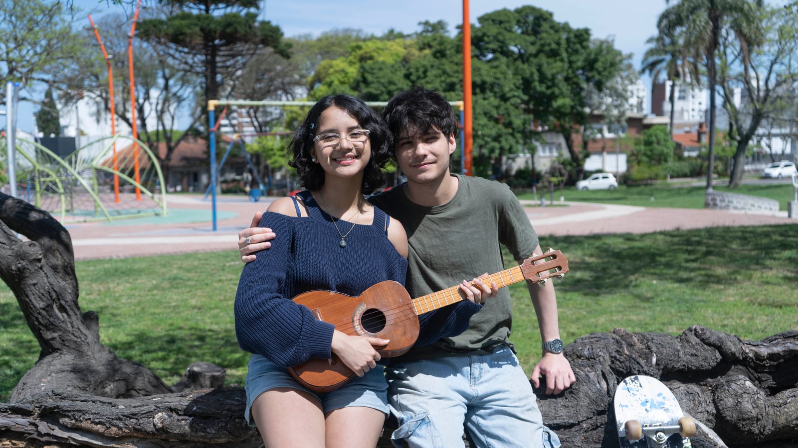 Una joven que sostiene un instrumento musical sentada junto a un joven en el tronco de un árbol en un parque