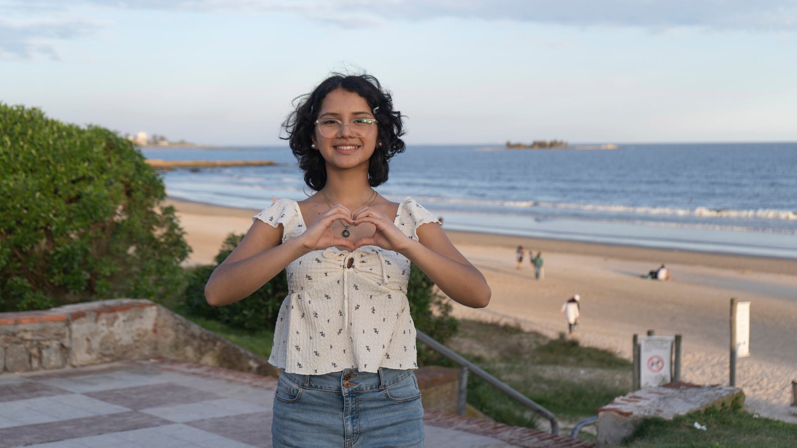 Frente al mar, una joven de cabello corto con anteojos forma un corazón con sus manos