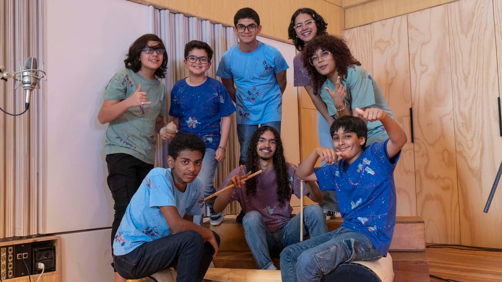 Un grupo de ocho adolescentes con playeras de colores y pantalones de mezclilla en un estudio de grabación
