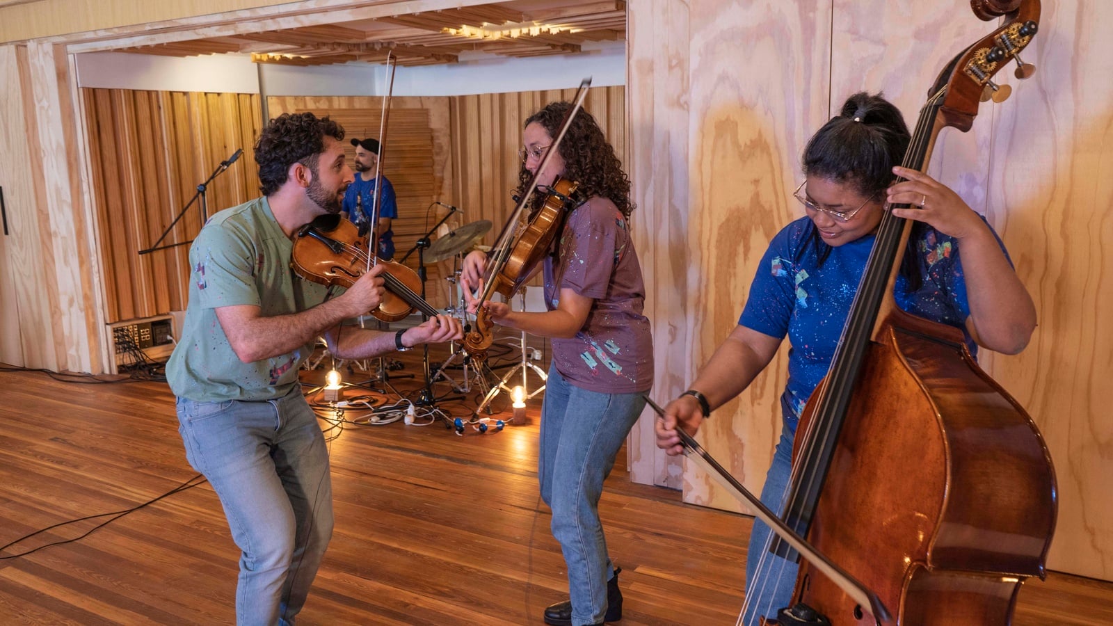 Tres personas de pie en un salón con techos, paredes y pisos de madera tocan diferentes instrumentos musicales 