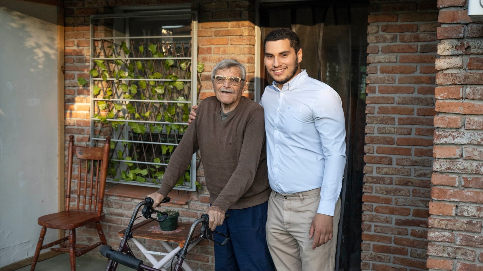 Un adulto mayor con andadera y un joven, posan al frente de una casa de ladrillo
