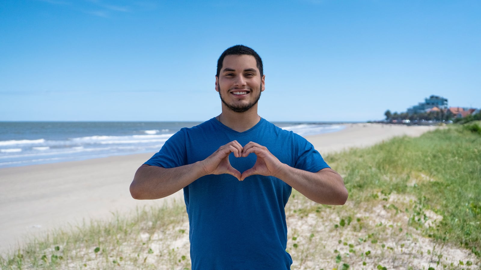 Un hombre joven que viste playera azul y está parado frente al mar, forma un corazón con sus manos