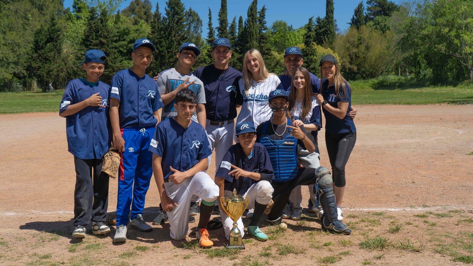 Ocho chicos y tres chicas vestidos con uniformes de béisbol azul marino, posan para la foto en un campo de juego