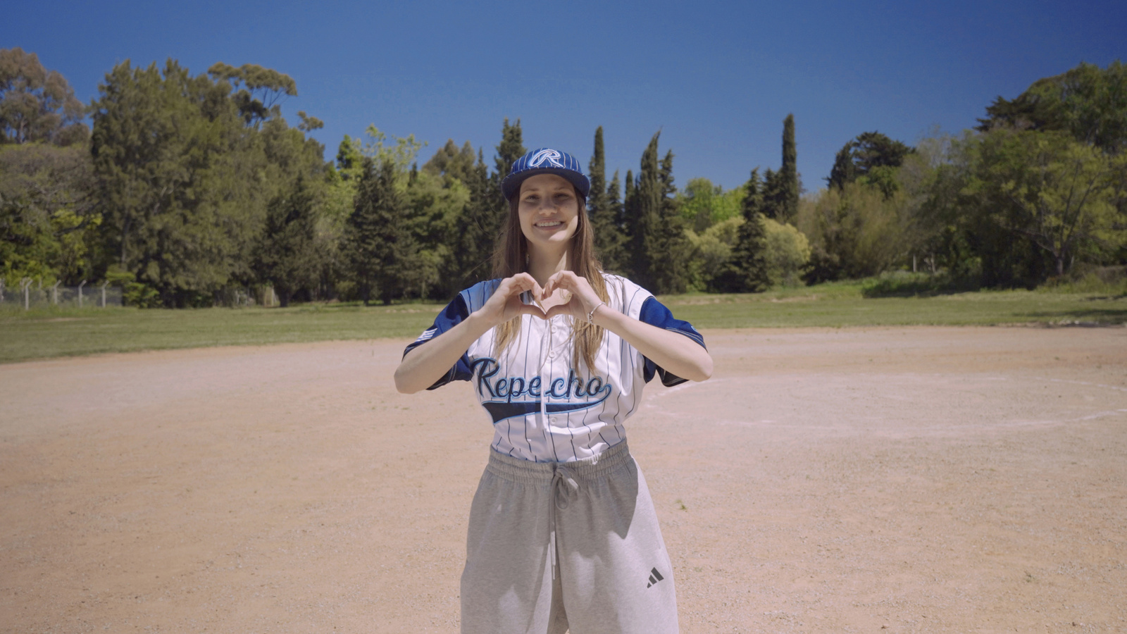 Una chica que viste uniforme de un equipo de béisbol, forma un corazón con sus manos en un campo de juego