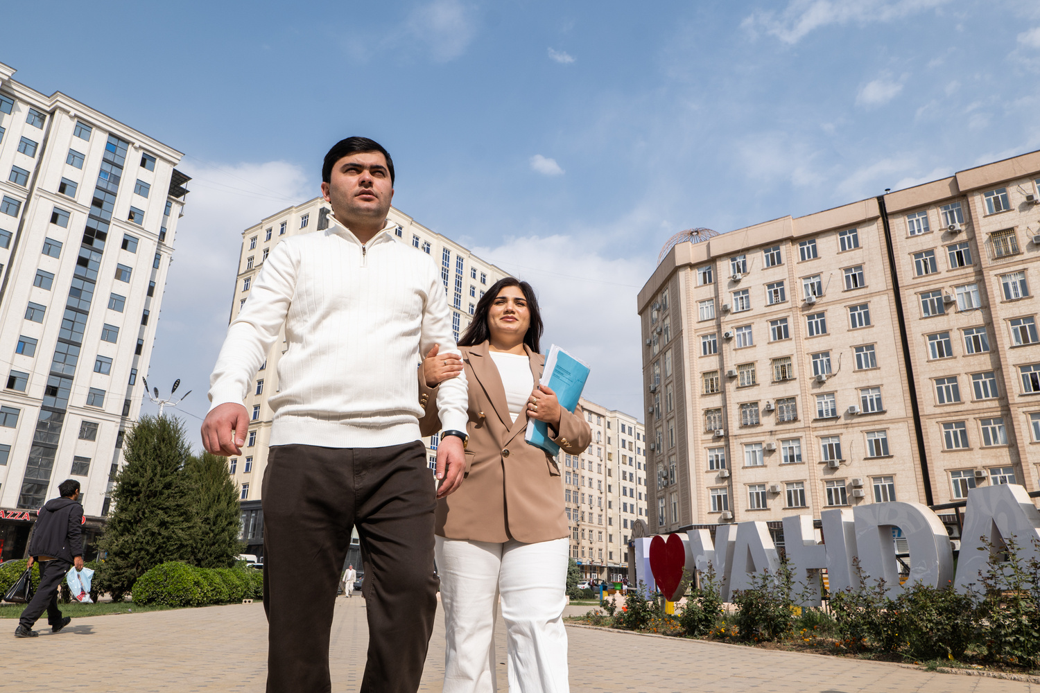 Una pareja camina tomada del brazo en una plaza rodeada de altos edificios de apartamentos