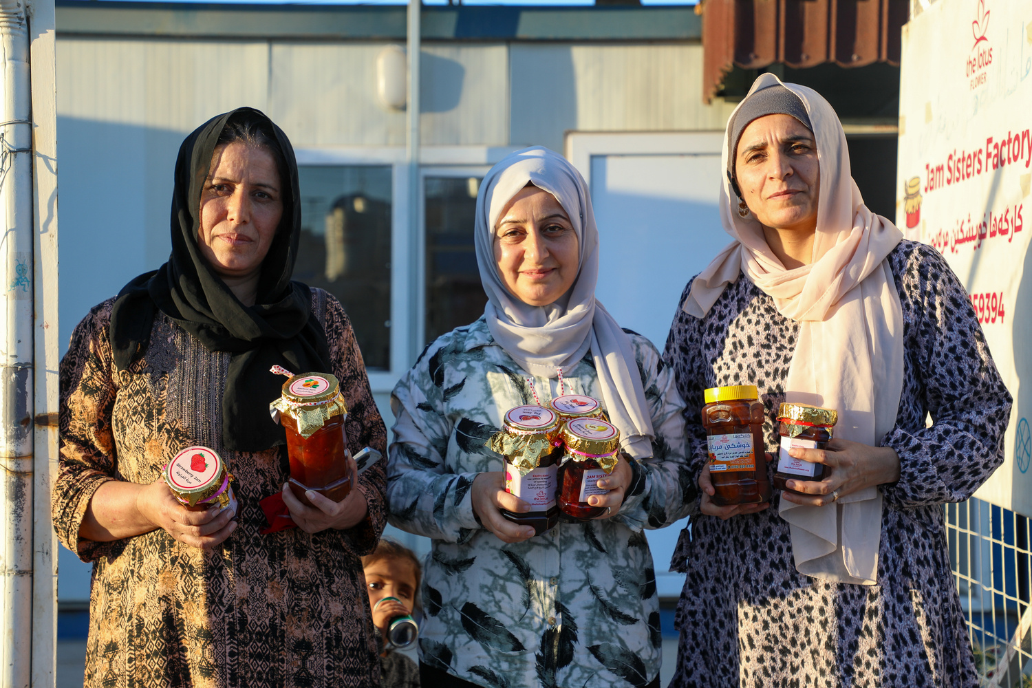 Tres mujeres posan frente a un edificio temporal a la luz del atardecer con tarros de mermelada casera en las manos