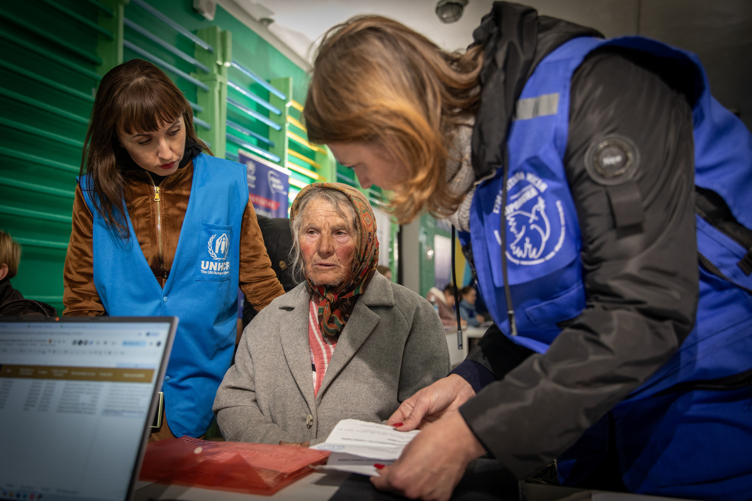 Dos trabajadoras humanitarias junto a una mujer mayor sentada frente a un escritorio