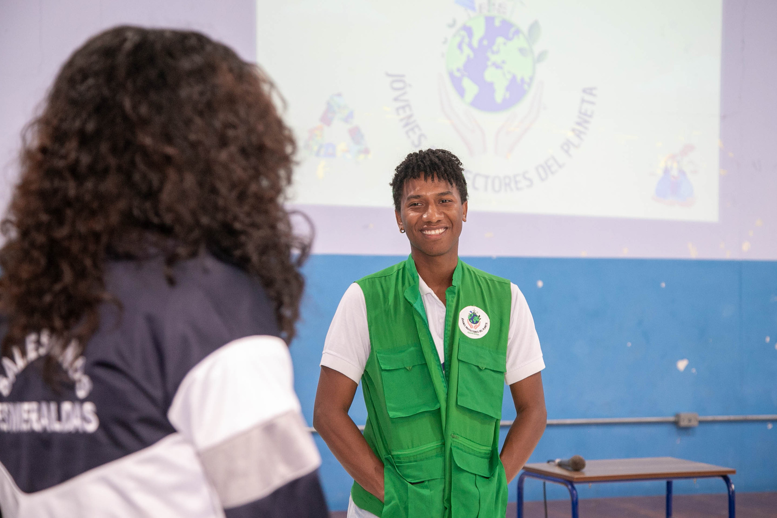 En segundo plano un joven con chaleco de verde sonríe hacia una mujer que de encuentra de espaldas en un aula