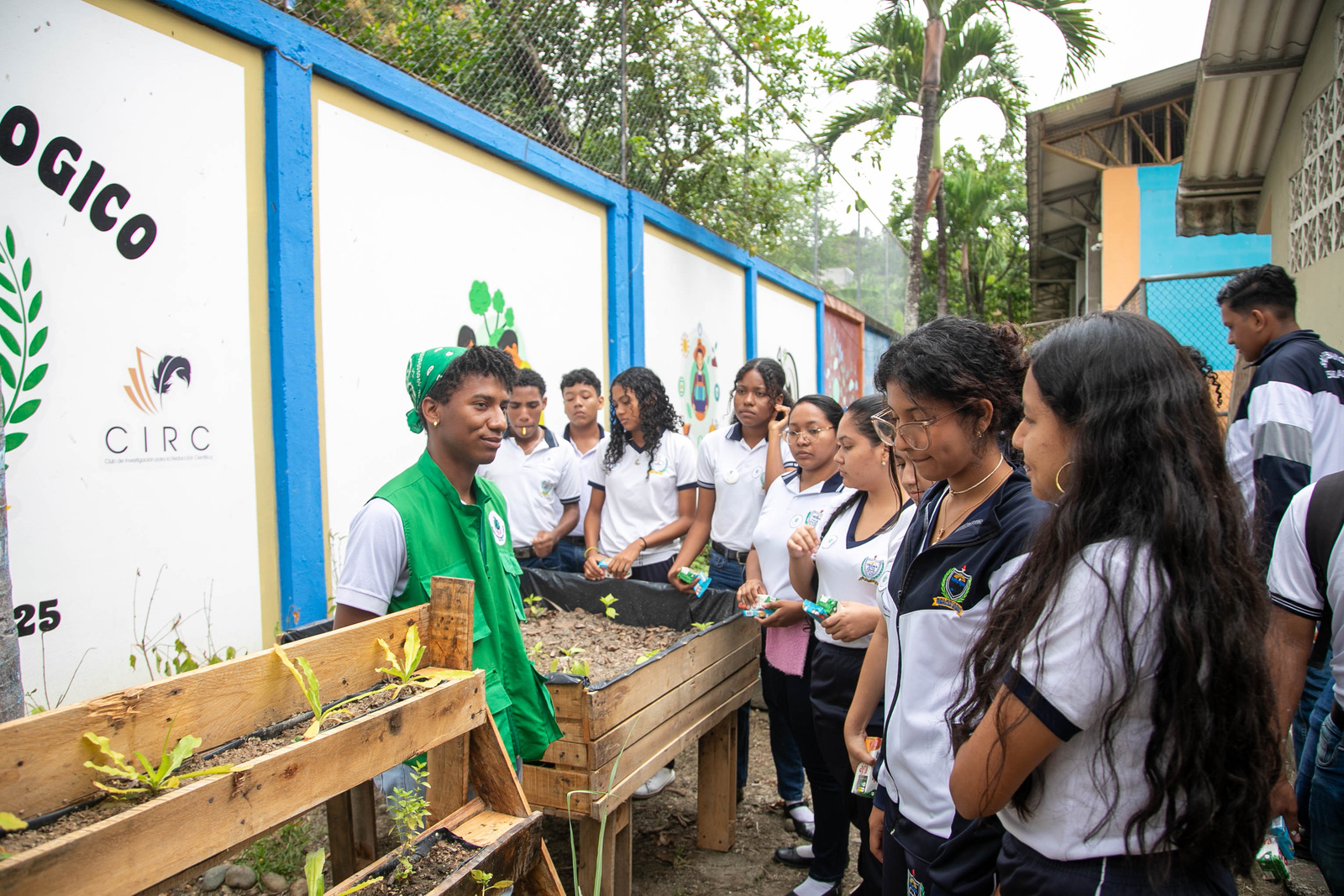 Un joven con chaleco verde junto a un grupo de estudiantes en un huerto
