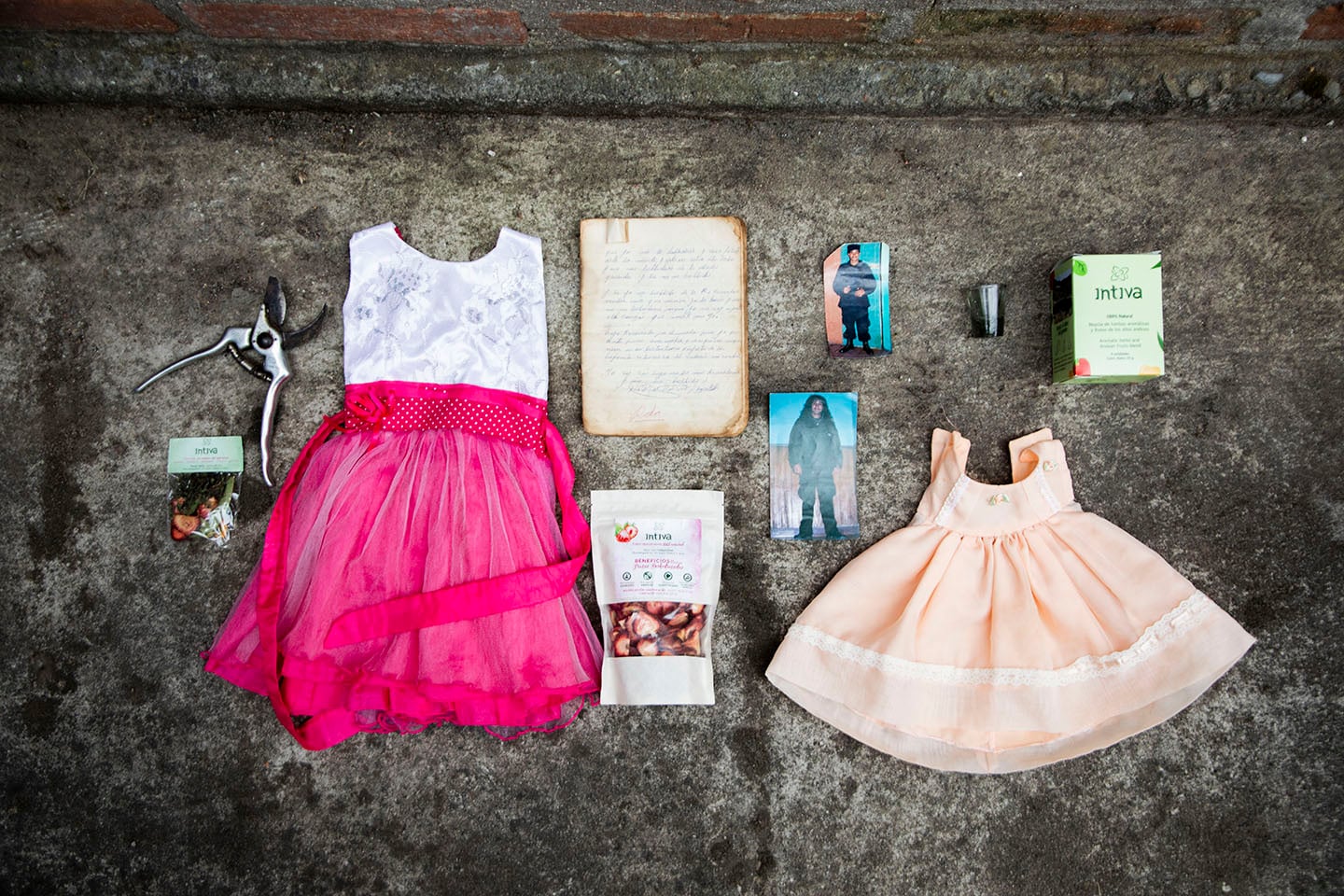 Objetos personales ordenados en una superficie plana: hay dos vestiditos de niña, unas fotos, páginas de cuaderno, y otros objetos.