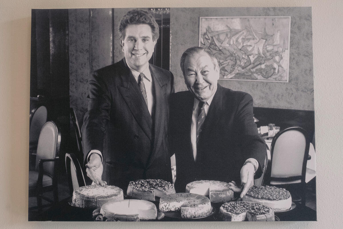 Fotografía en blanco y negro de dos hombres de pie detrás de una mesa llena de diferentes pasteles de queso.