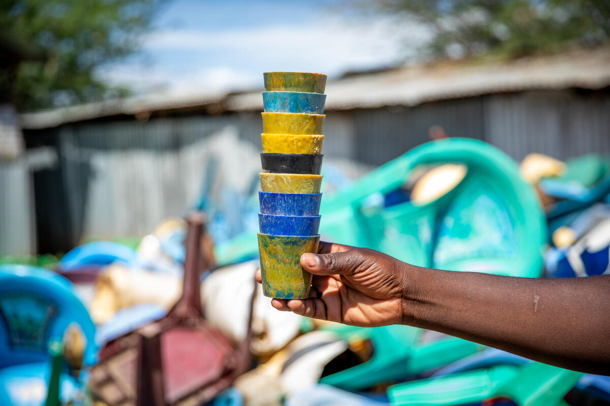 Una mano sostiene una pila de vasos sobrepuestos que están hechos de plástico reciclado; al fondo, un montón de residuos plásticos.