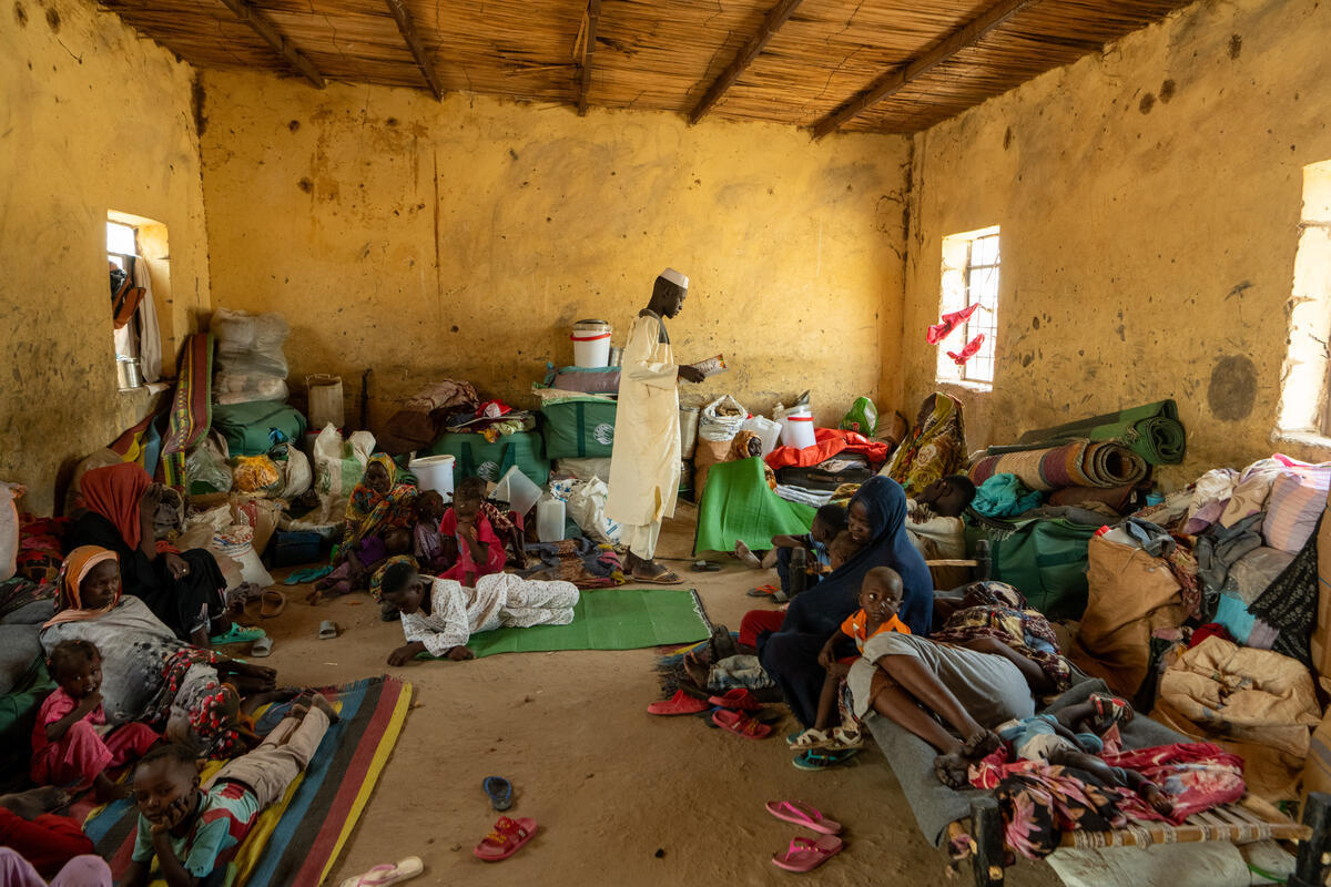 Un joven está leyendo mientras mujeres y niños están sentados o acostados en una habitación abarrotada de sus pertenencias