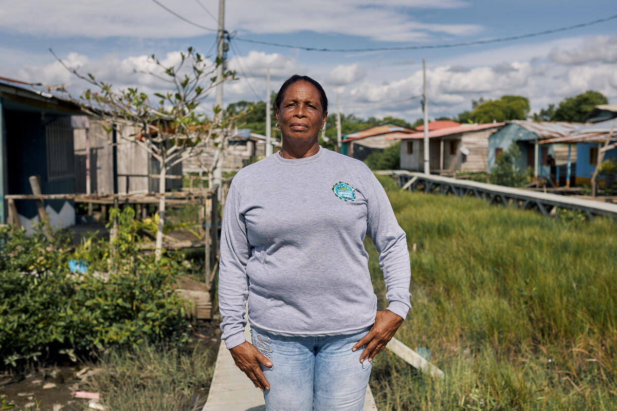 Una mujer afrocolombiana en medio de un terreno verde; detrás suyo, varias casas.