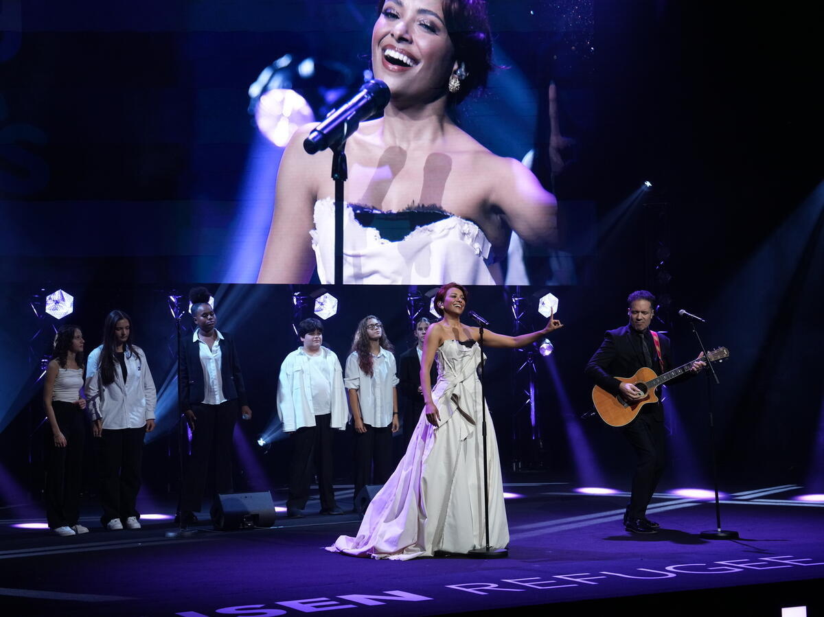 Una mujer en el escenario con un guitarrista y un coro juvenil delante de una pantalla gigante