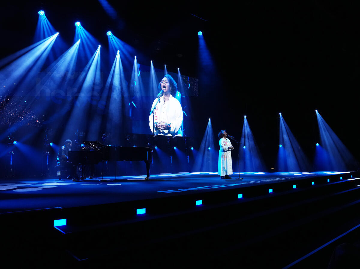 Una mujer vestida de blanco canta en el escenario respaldada por pianista y una pantalla gigante