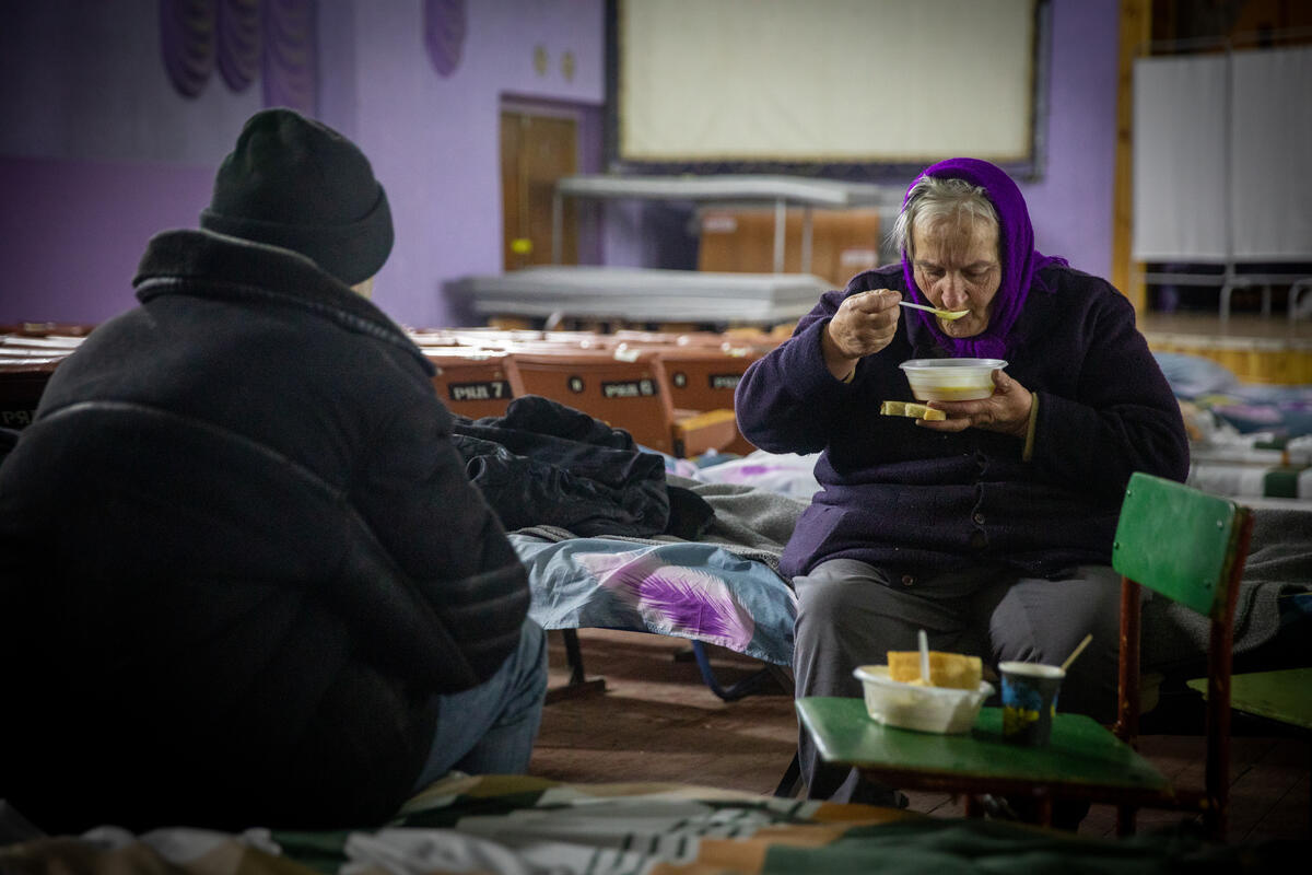 Sentada en una cama, una mujer mayor come mientras sostiene un plato hondo con la mano izquierda. Frente a ella, un hombre que lleva puesto un gorro y le da la espalda a la cámara.