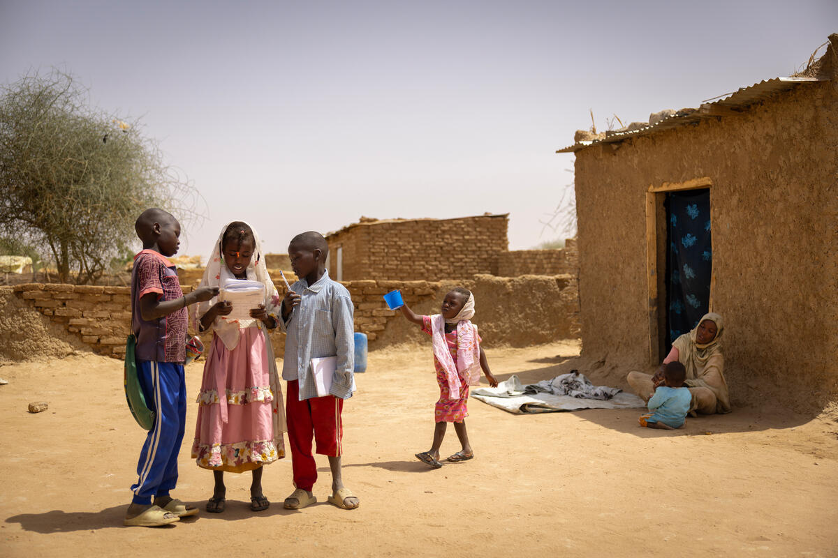 Cuatro niños posan junto a su madre y su hermano pequeño, que está sentado a la sombra frente a una pequeña casa de adobe.