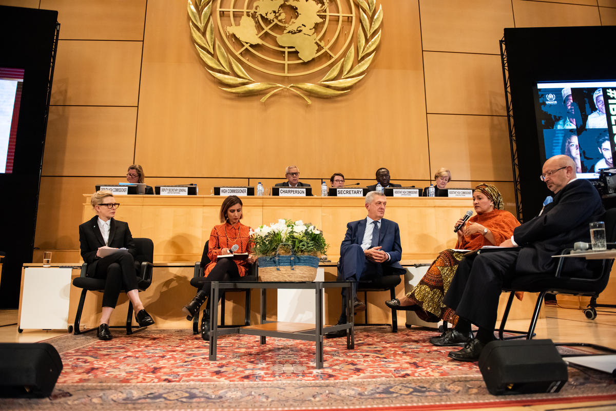 En un recinto de la ONU, un grupo de personas (dos hombres y tres mujeres) conversan en un semicírculo; detrás suyo, una hilera de personas sentadas detrás de una señalética que precisa el cargo de cada una. Hay dos lugares vacíos en el panel.
