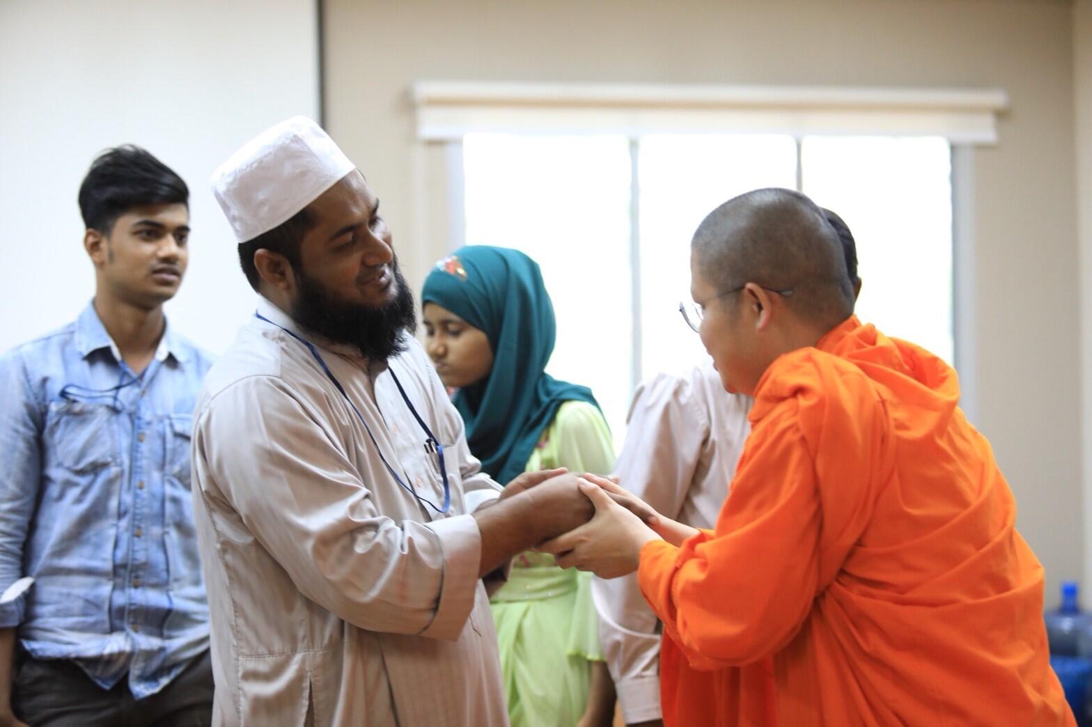 Un monje budista saluda a un hombre rohingya refugiado; detrás de ambos hay dos personas, un hombre y una mujer.