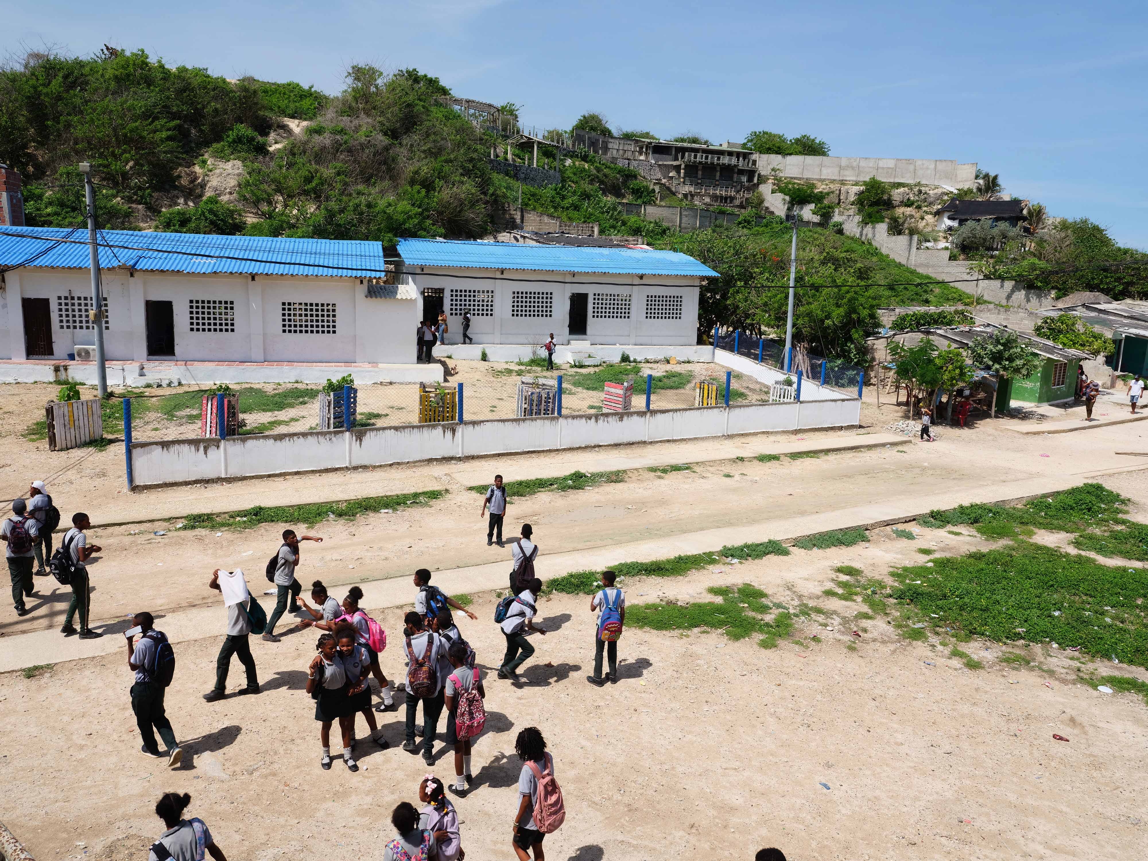 Alrededor de 30 estudiantes a la salida de una escuela. Visten uniformes y cargan sus mochilas.