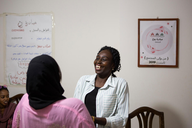 Dos mujeres conversan y ríen en una sala; una de ellas le está dando la espalda a la cámara y lleva puesto un hijab