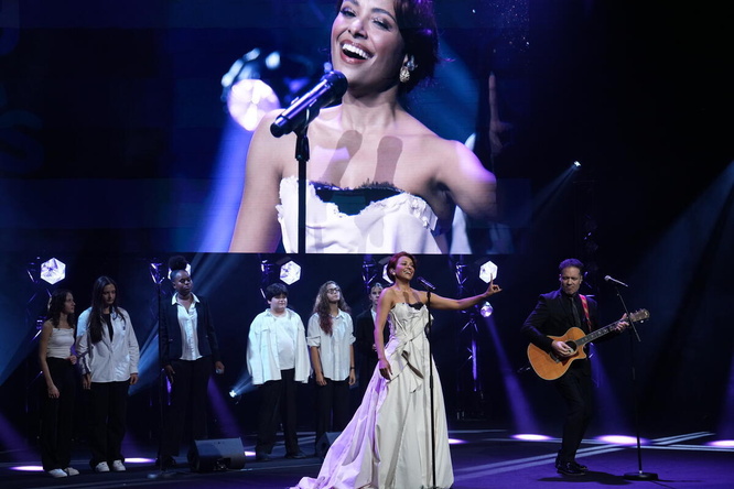 Una mujer en el escenario con un guitarrista y un coro juvenil delante de una pantalla gigante