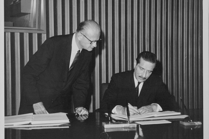Imagen en blanco y negro. Dos hombres (uno, sentado; el otro, de pie a su lado). El hombre sentado, que aparece a la derecha, está firmando un documento; el otro observa.