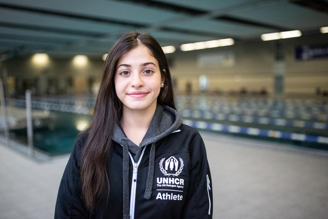 Una mujer joven que lleva puesta una chaqueta negra con el logotipo de ACNUR sonríe a la cámara; detrás suyo, una piscina.
