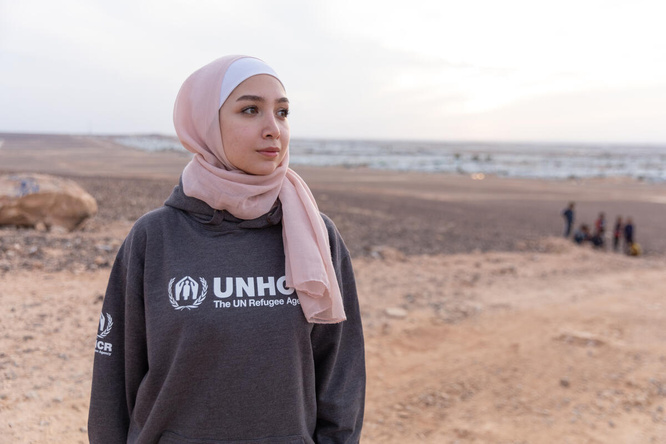 Una joven siria, que lleva puesto un hiyab rosado, mira al horizonte en medio de un terreno vacío; al fondo, un campamento de refugiados.