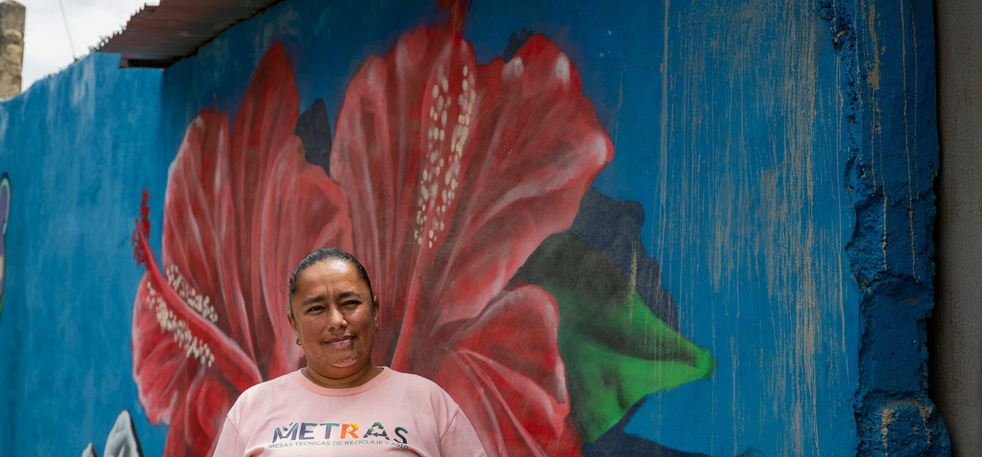 Una mujer con camiseta rosa, de pie frente a una pared azul que tiene pintada una flor roja grande con hojas verdes