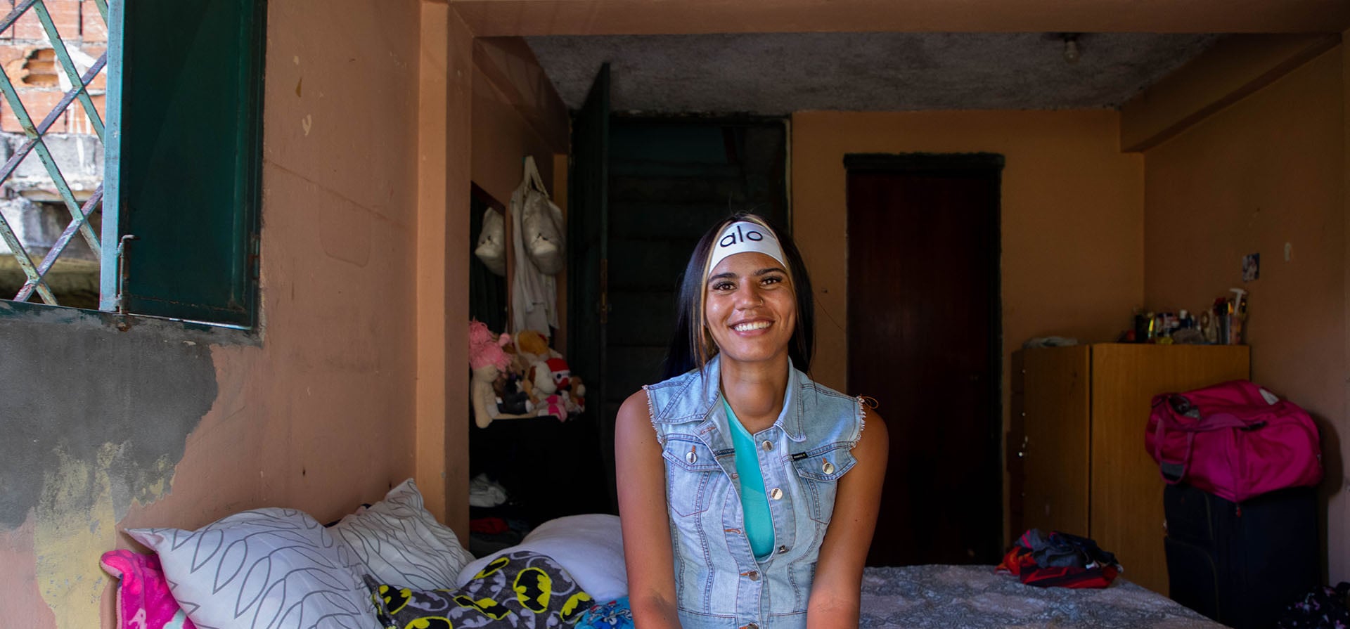 Una mujer joven posa sonriendo sentada al borde de una cama tendida en una habitación colorida.