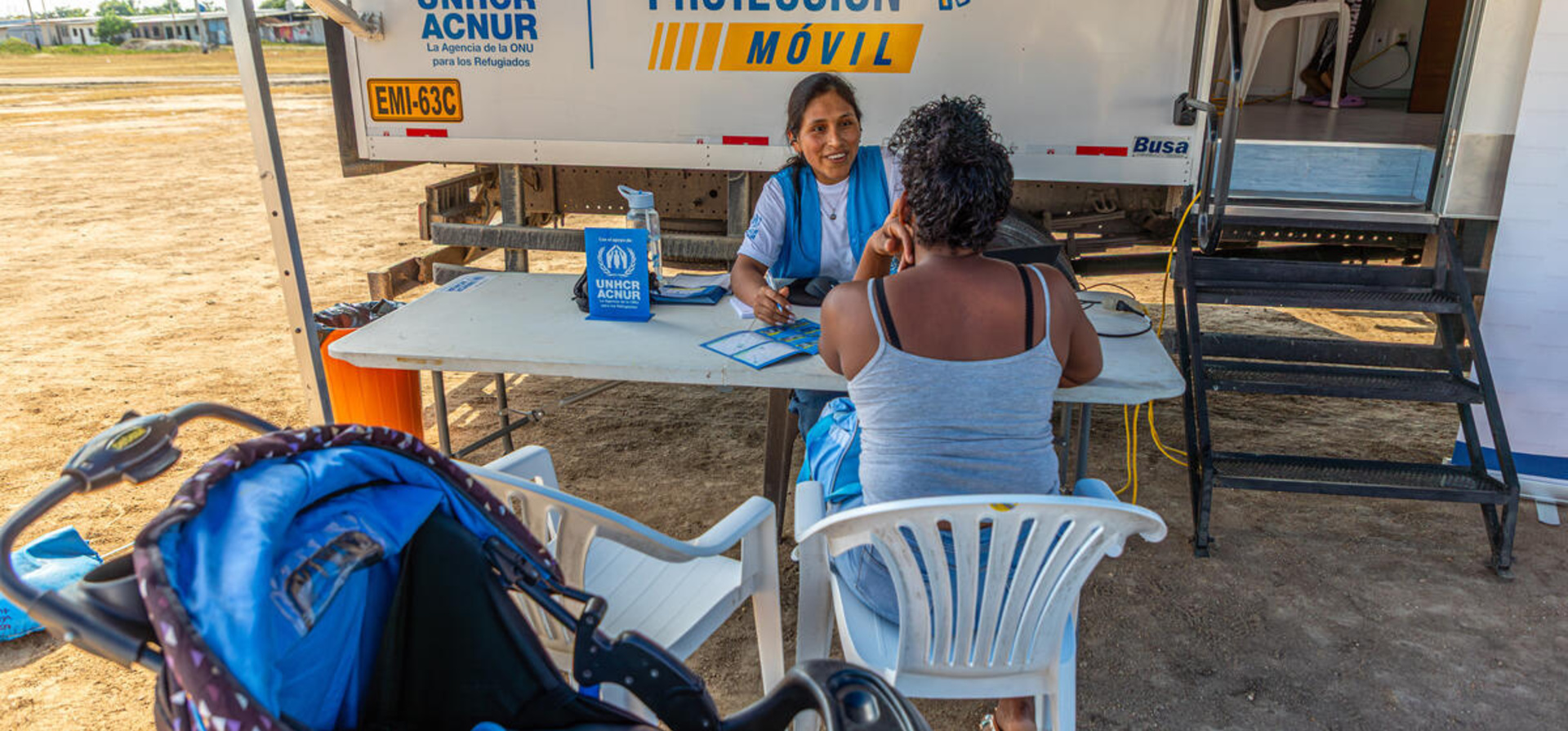 Una mujer con uniforme de ACNUR, camiseta blanca y chaleco azul, habla con una mujer que se encuentra de espaldas sentada en una silla plástica blanca
