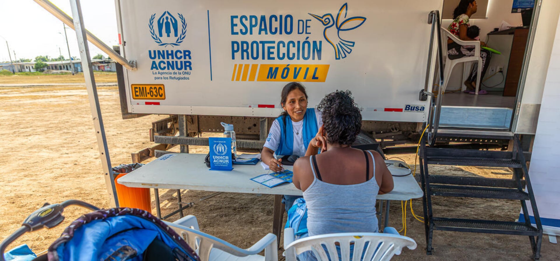 Una mujer con uniforme de ACNUR, camiseta blanca y chaleco azul, habla con una mujer que se encuentra de espaldas sentada en una silla plástica blanca