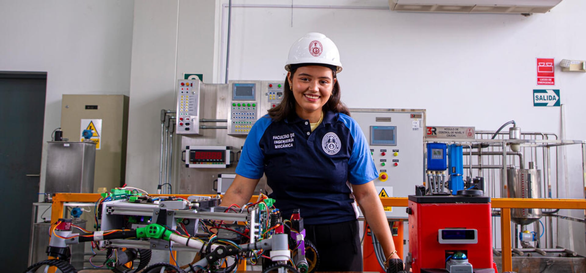 Yuliveth posa para una foto en un laboratorio rodeada dispositivos y mostrando en la mesa delante de ella varios modelos de pequeños robots.