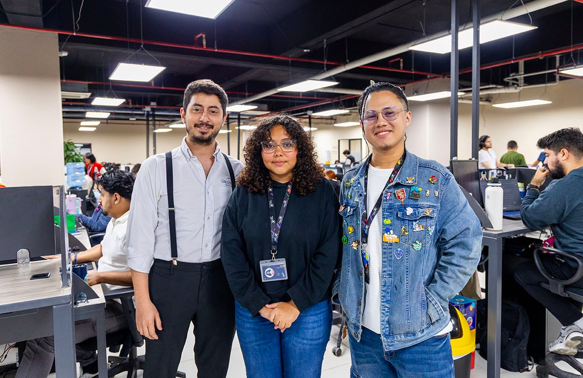 Dos hombres y una mujer posan juntos para una foto en una oficina abierta con escritorios, computadoras y personal trabajando.