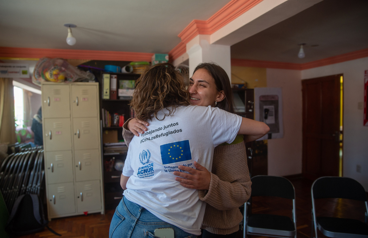 Dos mujeres se abrazan, una de ellas porta una playera blanca con logos de ACNUR y la Unión Europea