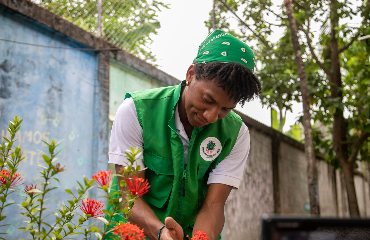 Un joven con un pañuelo verde en la cabeza y chaleco verde, toma entre sus manos unas flores en un jardín