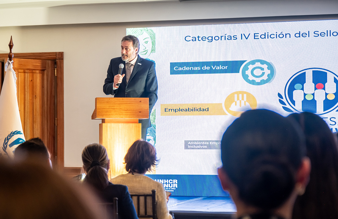 Un hombre de traje hablando desde un atril en un evento en un espacio cerrado con varios asistentes.
