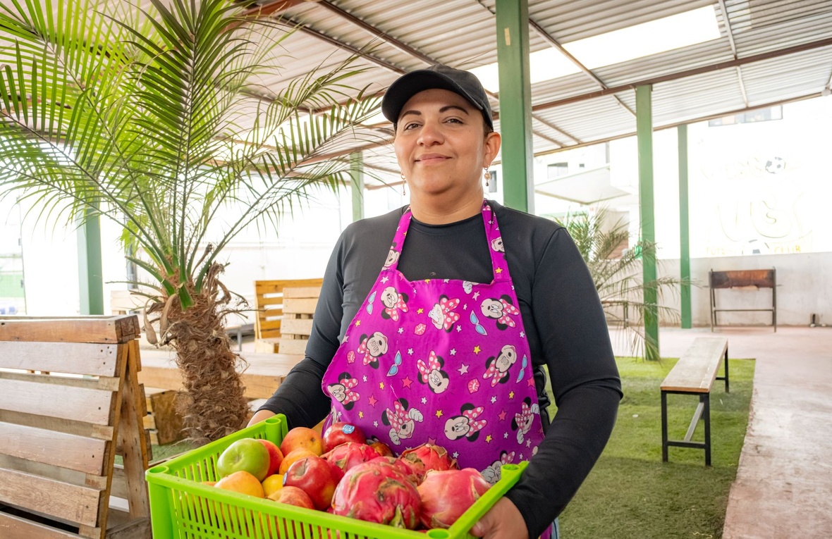 Una mujer sonríe a la cámara cargando una caja de frutas coloridas.