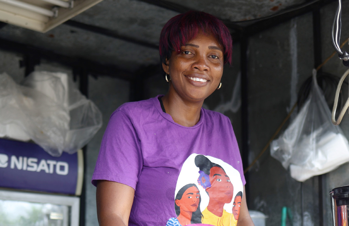 Una mujer con playera morada sonríe desde la ventana de un comedor comunitario