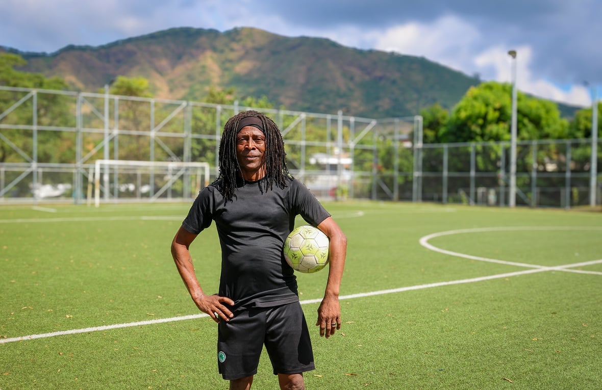 Un hombre de piel oscura y cabello largo oscuro vestido de negro, posa para la foto en una cancha de fútbol con pasto verde mientras sostiene con su brazo izquierdo un balón de fútbol