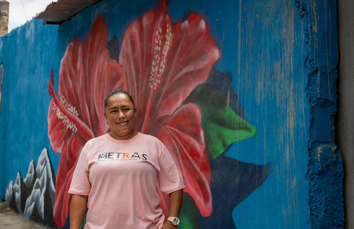 Una mujer con camiseta rosa, de pie frente a una pared azul que tiene pintada una flor roja grande con hojas verdes