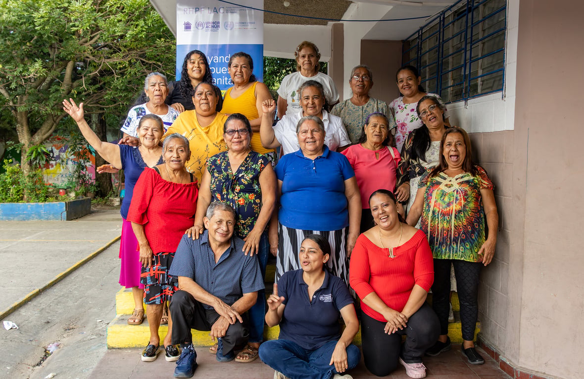 Quince mujeres adultas mayores, dos mujeres jóvenes, y un hombre adulto mayor, posan para la foto a las afueras de un aula